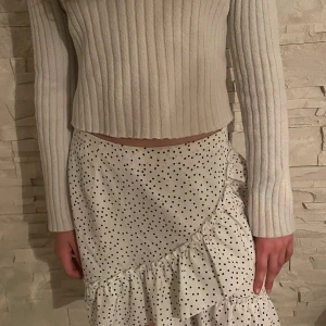 Vit prickig kjol med volang - Säljer en vit kjol med svarta prickar och volangdetalj längs nederkanten. Kjolen har omlottkänsla och stängs med dragkedja bak. Perfekt för dig som gillar en feminin och trendig stil. Pris kan diskuteras.