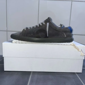 Oliver Cabell - Säljer ett par stilrena grå sneakers från Oliver Cabell i mocka med vita snören och diskreta detaljer. Skorna har en klassisk rund tå och platt sula, perfekt för en clean och avslappnad look.