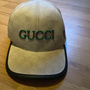 Svart  keps från Gucci - Snygg beige keps från Gucci med broderad logga framtill i grönt och rött. Kepsen har klassiskt mönster, svarta detaljer på skärmen och ett band i grönt och rött på sidan. Tillverkad i 50% bomull och 50% polyester.