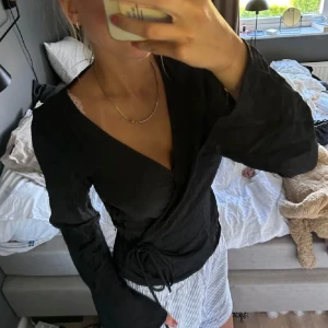 Svart omlottopp med långa ärmar - Säljer en svart omlottopp med långa ärmar och knyt i midjan. Toppen har en djup v-ringning och är tillverkad i ett mjukt tyg. Perfekt för att styla med både jeans och kjol.