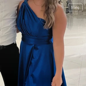 Blå långklänning med oneshoulder - Säljer en elegant blå långklänning med oneshoulder och markerad midja. Klänningen har en vacker glans och ett brett band i midjan samt slits framtill. Perfekt för bal eller fest!