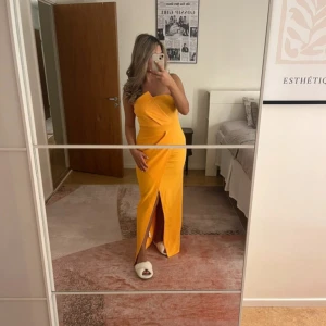 Gul långklänning med slits från Boohoo - Säljer en snygg gul långklänning från Boohoo med oneshoulder och hög slits framtill. Klänningen har figurnära passform och är tillverkad i ett slätt, stretchigt material. Perfekt för dig som vill sticka ut med en färgstark look!