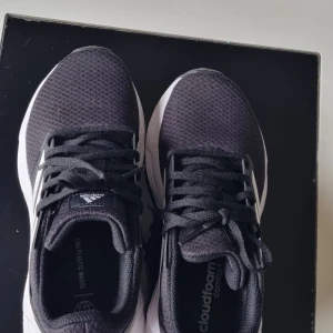 Svarta Adidas sneakers med vit sula - Säljer ett par svarta Adidas sneakers med vit sula och klassiska vita ränder på sidan. Skorna har snörning och är tillverkade i syntetmaterial med Cloudfoam Comfort-innersula för extra komfort. Perfekta för vardagsbruk och träning.