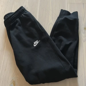 Svarta mjukisbyxor från Nike - Svarta mjukisbyxor från Nike med broderad vit logga på benet. Byxorna har resår i midjan och är tillverkade i mjukt bomullsmaterial. Perfekta för en avslappnad stil. Nya, Storlek: S/M