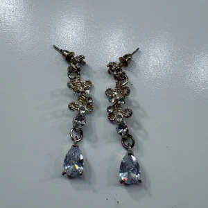 Glittriga droppformade örhängen - Säljer ett par eleganta örhängen i silverfärg med gnistrande stenar och droppformad kristall längst ner. Perfekta för att lyfta din look med extra glamour och stil.