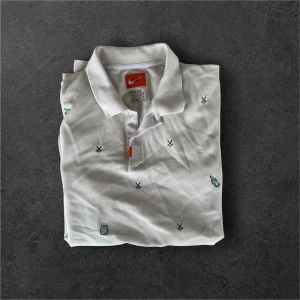 Vit golf pikétröja från Nike - Säljer en vit pikétröja från Nike med små broderade detaljer i grönt och svart. Tröjan har klassisk krage och korta ärmar, tillverkad i ett lätt och svalt material med Dri-Fit-teknologi.
