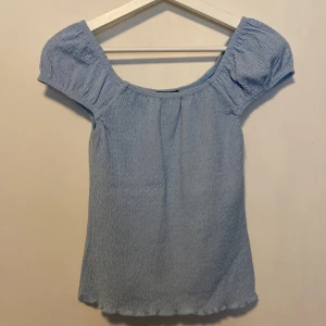 Ljusblå topp SisterS Point - Supersöt ljusblå topp från SisterS Point med puffiga korta ärmar och smockat tyg. Toppen har en rundad halsringning och är lätt stretchig för en bekväm passform. Perfekt för vår och sommar!