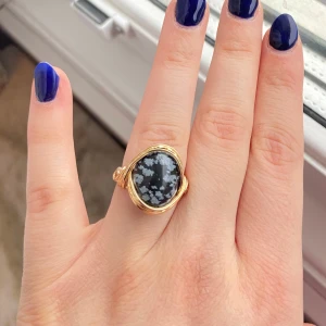 Guldfärgad ring med snöflingeobsidian - Unik ring med en stor oval snöflingeobsidian infattad i guldfärgad metall. Stenen har ett svart och grått mönster som påminner om snöflingor. Perfekt statement-smycke för dig som gillar naturstenar och iögonfallande accessoarer.