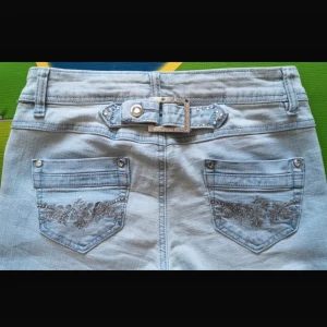 Ljusblå jeansbyxor med broderade bakfickor och bälte - Säljer ett par ljusblå jeansbyxor med snygga broderier på bakfickorna och ett dekorativt bälte med nitar och spänne. Byxorna har en rak passform och är tillverkade i klassiskt denimtyg. Perfekta för dig som gillar detaljer och vill sticka ut lite extra.