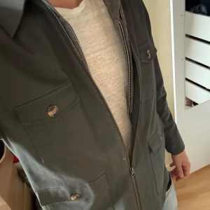 Mörkgrön John Henric jacka - Säljer en mörkgrön jacka med dragkedja framtill och fyra fickor med knappar. Klassisk krage och rak passform. Perfekt för dig som vill ha en stilren och enkel jacka till vardags.
