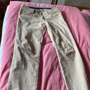 Beige chinos från Hansen & Jacob - Snygga beige chinos från Hansen & Jacob med klassisk rak passform. Byxorna har fem fickor, diskreta detaljer i grönt och blått samt en liten metallogo framtill. Tillverkade i bomull med lite stretch för extra komfort.