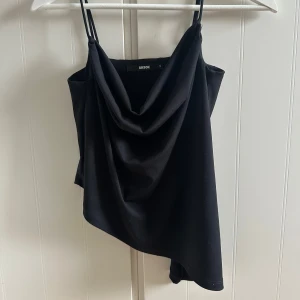 Svart asymmetrisk linnetopp från BikBok - Säljer en svart linnetopp från BikBok med tunna axelband och snygg draperad halsringning. Toppen har en asymmetrisk nederkant som ger en cool look. Perfekt att styla till olika outfits!