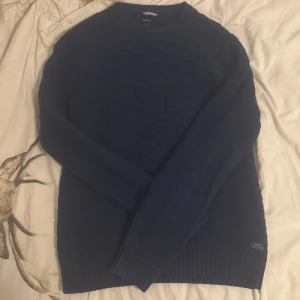 Mörkblå stickad tröja från GANT - Säljer en mörkblå stickad tröja från GANT i storlek M. Tröjan har ett diskret rutmönster, rund halsringning och ribbade muddar vid ärmslut och nederkant. Perfekt för kyligare dagar och nu till sommaren. 