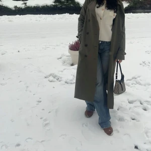 Olivgrön trenchcoat  - Säljer en olivgrön trenchcoat  med klassisk krage och knappar framtill. Jackan är lång och har en avslappnad passform, perfekt att slänga över en outfit för en cool look.
