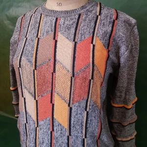 Mönstrad stickad tröja i retrostil - Säljer en gråmelerad stickad tröja med färgglada geometriska mönster i orange, beige, gul och röd. Tröjan har rund halsringning och långa ärmar med dekorativa ränder. Perfekt för dig som gillar vintage och unika plagg.