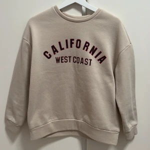 Beige sweatshirt med tryck från SHEIN - Beige sweatshirt från SHEIN med texten 'California West Coast' i rött och svart framtill. Tröjan har rund halsringning och långa ärmar. Perfekt för en avslappnad och trendig stil.