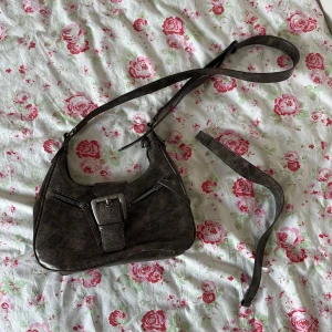 Brun väska - Säljer en brun väska axelband och stort dekorativt spänne framtill. Väskan har dragkedja upptill och innerficka. Perfekt för att bära det viktigaste och ger en cool vintagekänsla.