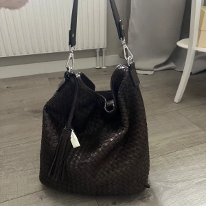 Mörkbrun axelväska från Suri Frey - Säljer en mörkbrun axelväska från Suri Frey med flätad design och dekorativ tofs. Väskan har silverfärgade metalldetaljer och dragkedja upptill. Perfekt storlek för vardags eller skolan och har en avtagbar axelrem. Den har en liten defekt på kanten där en fläta släppt med det går lätt att åtgärda.
