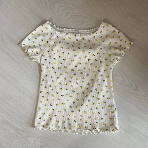 Vit ribbad topp med citronmönster från Lindex - Superfin vit ribbad topp från Lindex med små gula citroner över hela. Toppen har korta ärmar och vågig kant vid hals, ärmslut och nederkant. Perfekt för dig som gillar färgglada och lekfulla plagg!