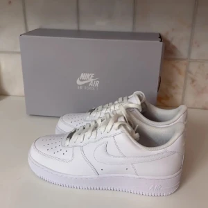 Nike Air Force 1 vita sneakers - Klassiska helvita Nike Air Force 1 sneakers med låg profil och perforerad tå. Skorna har snörning och är tillverkade i skinn med en platt sula. Perfekta för en clean och stilren look.