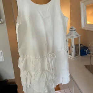 Vit volangklänning med knytband - Säljer en söt vit klänning med volanger och knytband i midjan. Klänningen är ärmlös och har en rund halsringning. Perfekt för sommarens alla tillfällen! Den är helt oanvänd och prislappen sitter kvar. Hör av vid frågor 😊