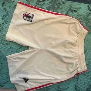 Vit-beiga Adidas shorts från Japans fotbollslandslag, stl M - Säljer ett par vita-beiga Adidas shorts med röda ränder längs sidorna och klubbmärke på ena benet. Klassisk sportig look med resår i midjan. Perfekta för fotboll eller träning.