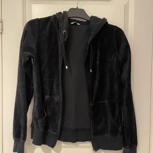Svart hoodiejacka i velour - Mjuk och skön svart hoodiejacka i velour med dragkedja framtill och två fickor. Jackan har huva med snören och ribbade muddar vid ärmslut och nederkant. Perfekt för en avslappnad stil.