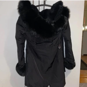 Svart parkajacka med äkta rävpäls  - Säljer en svart parkajacka med stor huva och breda pälsdetaljer runt huva och ärmslut. Jackan har dragkedja och knappar framtill samt en längre passform. Perfekt för kalla dagar.