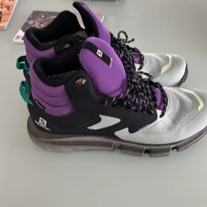 Salomon sneakers i lila och svart - Säljer ett par Salomon sneakers i lila, svart och grått med gröna detaljer. Skorna har snörning, hög modell och robust sula. Perfekta för dig som gillar streetwear eller outdoor-stil.