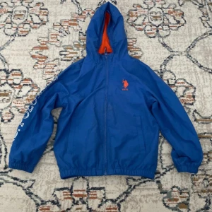 Blå vindjacka med huva från U.S. Polo Assn. - Blå vindjacka med huva och dragkedja från U.S. Polo Assn. Jackan har orange innerfoder och tryck på bröstet samt ryggen. Elastiska muddar vid ärmslut och nederkant. Perfekt för blåsiga dagar.