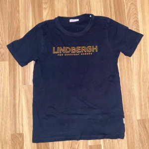 Svart t-shirt från Lindbergh - Svart t-shirt från Lindbergh med tryckt text i orange på bröstet. Klassisk rund halsringning och korta ärmar. Perfekt för en avslappnad stil.
