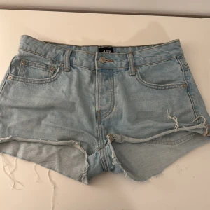 Ljusblå jeansshorts med från 157 - Säljer ett par ljusblå jeansshorts från 157 med råa fransade kanter och klassisk femficksdesign. Shortsen har knappgylf och bälteshällor, perfekt för en avslappnad stil.