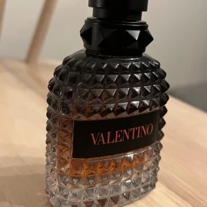 Valentino parfymflaska med nitar - Säljer en lyxig parfym från Valentino i en cool, nitprydd glasflaska med svart och transparent design. Flaskan har en svart etikett med röd text och innehållet är bärnstensfärgat. Perfekt för dig som gillar exklusiva dofter och snygg design.