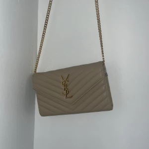 Beige quiltad axelväska från YSL - Snygg beige axelväska från YSL med guldfärgad kedja och ikonisk YSL-logga framtill. Väskan har quiltad design i chevronmönster och är tillverkad i skinn. Perfekt för dig som vill ha en stilren och lyxig väska med exklusiv känsla.
