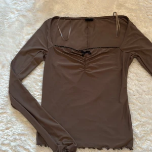 Brun långärmad topp från Gina Tricot - Säljer en brun långärmad topp från Gina Tricot med söt svart rosett och rynkdetalj framtill. Toppen har smala axelband, svarta dekorativa kanter och är figurnära i modellen. Aldrig använd helt nytt skick