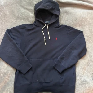 Mörkblå hoodie från Polo Ralph Lauren - Säljer en mörkblå hoodie från Polo Ralph Lauren med klassisk röd logga på bröstet. Tröjan har huva med vita snören, känguruficka och är tillverkad i mjuk bomull. Perfekt för chill dagar och enkel att matcha med jeans eller joggers. Tröjan har inga defekter och är i toppen skick. Ny pris: 2000kr Mitt pris: 900kr. Storlek Medium Herr