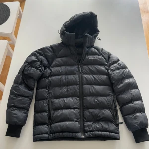 Svart dunjacka från Peak Performance - Snygg svart dunjacka från Peak Performance med huva och dragkedja framtill. Storlek 170