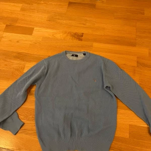 Ljusblå stickad tröja från Gant - Snygg ljusblå stickad tröja från Gant med rund halsringning och diskret logga broderad på bröstet. Tröjan har ribbade muddar vid ärmslut och nederkant, perfekt för en stilren och avslappnad look.