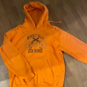 Orange hoodie från Menace, strl L - Snygg orange hoodie från Menace med tryck 'Beverly Hills Gun Range' och text på ärmen. Klassisk känguruficka, snörning i huvan och skön bomullskvalitet. Perfekt för dig som gillar streetwear och vill sticka ut med färg.
