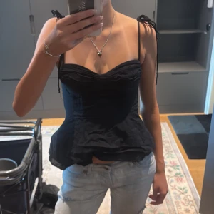 Svart korsett-topp med knytband - Supersnygg svart korsett-topp med smickrande sweetheart-urringning och knytbara axelband. Toppen har figurnära passform och lätt utsvängd nederdel som ger en cool siluett. Perfekt att styla med jeans eller kjol för en trendig look.