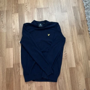 Mörkblå stickad tröja från Lyle & Scott - Snygg mörkblå stickad tröja från Lyle & Scott med klassisk rund hals och gul broderad logga på bröstet. Tröjan har långa ärmar och är tillverkad i mjukt material som passar perfekt till lager-på-lager-stil.