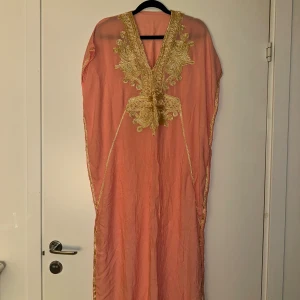Korallfärgad långklänning med guldmönster - Säljer en korallfärgad långklänning med v-ringning och vackra broderade gulddetaljer framtill och längs kanterna. Klänningen har lös passform och är perfekt för dig som vill sticka ut med eleganta detaljer och unik stil.