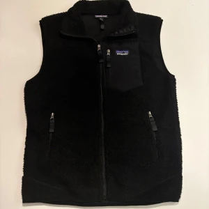 Svart fleeceväst från Patagonia - Svart fleeceväst från Patagonia med hög krage och hel dragkedja framtill. Västen har tre fickor med dragkedja, varav en på bröstet, och ett mjukt, fluffigt material som håller dig varm. Perfekt för lager-på-lager och outdoor-vibes.