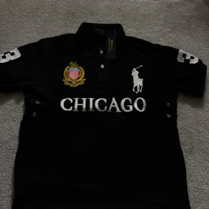 Chief Keef Polo  - Svart t-shirt från Polo Ralph Lauren med broderad logga och emblem på bröstet, samt stor vit text 'CHICAGO' framtill. Slim fit-modell med korta ärmar och klassisk krage med knappar. Snygga vita detaljer på ärmarna och broderade inslag i guld och vitt. Perfekt för dig som gillar sportig stil. Helt ny tröja, helt oanvänd! DM:A om ni har fler frågor 