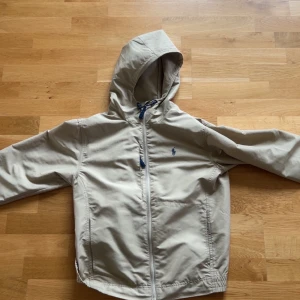 Beige vindjacka från Polo Ralph Lauren - Snygg beige vindjacka från Polo Ralph Lauren med huva och dragsko. Jackan har dragkedja framtill, två sidofickor och en broderad logga på bröstet. Perfekt för blåsiga dagar och enkel att matcha med det mesta.