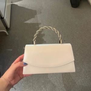 Vit kuvertväska med flätat handtag - Stilren vit kuvertväska med snyggt flätat handtag i silverfärgad metall. 