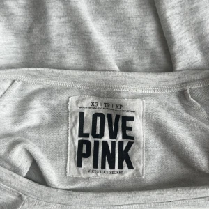 Grå sweatshirt från Victoria's Secret PINK - Ljusgrå sweatshirt från Victoria's Secret PINK med svart tryck 'LOVE PINK LOVE' på bröstet. Tröjan har rn off shoulder halsringning och långa ärmar. Mjuk och skön i bomullsblandning, perfekt för chill dagar eller när du vill ha en enkel och snygg look.