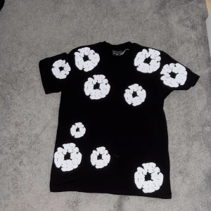 Denim tears t-shirt -  svart t-shirt med stora vita blommönster över hela plagget. Klassisk passform med korta ärmar och rund hals. Perfekt för dig som vill sticka ut med en unik och trendig look. Materialet känns mjukt och bekvämt mot huden. Storlek är ungefär mellan M och S 