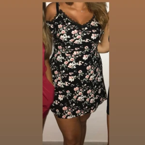 Svart blommig kort klänning - Säljer en svart kort klänning med rosa och vita blommor. Klänningen har offshoulder och axelband, samt spetsdetaljer vid halsringningen. Passformen är normal och tyget är mjukt och följsamt, perfekt för sommarens alla tillfällen. Använd endast en gång. 