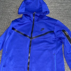 Blå Nike vindjacka med huva, Pris kan diskuteras!! - Nike vindjacka i stark blå färg med svart logga på bröstet och svarta detaljer längs dragkedjan och ärmen. Jackan har huva, hel dragkedja och lång ärm. Materialet är mjukt och syntetiskt, perfekt för att hålla vinden ute. Snygg sportig look med stilrena linjer.